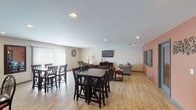 Located at Fountains Blvd in West Chester, Ohio, this single-story community space is designed to foster comfort and social interaction through a well-planned, open-concept layout. Light wood-tone flooring runs throughout the main areas, lending a warm, cohesive feel to the entire space.

The kitchen is outfitted with dark espresso cabinetry, stainless steel appliances including a refrigerator, range, and microwave, and a dark granite-topped island with bar seating. Pendant lighting above the island adds a modern accent, while tile flooring in the cooking area provides a practical transition underfoot.

The kitchen opens directly into a generous dining area furnished with multiple high-top tables and ladder-back chairs, accommodating a comfortable number of guests. Vertical blinds on the windows allow for flexible light control throughout the day.

Adjacent to the dining area, a spacious lounge features recessed lighting, a wall-mounted flat-screen television, and an arrangement of sofas and accent chairs suited for relaxed gathering. A distinctive terracotta-toned accent wall adds visual warmth to this zone, complemented by decorative wall art and a pass-through window that connects to neighboring areas. The overall ambiance is inviting, functional, and thoughtfully arranged for communal use.