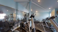 This fitness center offers a well-appointed workout environment designed to support a full range of exercise routines. The space is characterized by striking vaulted ceilings with exposed white beams that lend an open, airy feel, complemented by soft blue walls that create a calm and motivating atmosphere. Recessed lighting ensures the room is well-lit throughout.

The floor plan accommodates a diverse selection of exercise equipment, including elliptical trainers, treadmills, and a multi-station strength training machine, providing options for both cardiovascular and resistance training workouts. Dark hardwood-style flooring adds a polished, durable surface underfoot.

A large floor-to-ceiling mirror spans one wall, visually expanding the space while allowing users to monitor their form during exercise. The overall layout is thoughtfully organized to allow comfortable movement between equipment. Natural light filters in through a window near the entrance, further brightening the interior. The combination of architectural character, practical amenities, and a clean, inviting design makes this fitness center a functional and pleasant space for daily exercise.