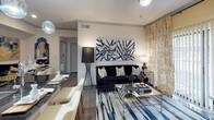 This single-floor residence showcases a cohesive, design-forward aesthetic anchored by a navy, gold, and black color palette. The open living and dining area features hardwood-style flooring, a velvet sofa, glass coffee table, and bold abstract artwork, while floor-to-ceiling gold-patterned drapes frame a sliding glass door that brings in natural light. Pendant lighting adds a contemporary touch overhead.

The galley-style kitchen is appointed with dark cabinetry, granite-style countertops, a mosaic tile backsplash, and stainless steel appliances, including a French door refrigerator and electric range. A doorway leads to an in-unit laundry area beyond.

The bedroom is a dramatic focal point, featuring a bold marbled-pattern accent wall in deep navy and grey behind an upholstered headboard. Soft carpet, dark floor-length curtains, natural window light, and a walk-in closet with built-in wire shelving and woven storage baskets complete the room.

The bathroom offers a clean, modern finish with dark-framed cabinetry, a granite-topped vanity, a large framed mirror, and contemporary fixtures, maintaining the unit's refined, monochromatic design throughout.