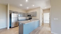 This well-appointed single-story home offers a thoughtfully designed layout that balances modern aesthetics with everyday functionality. The heart of the home is an open-concept kitchen and living area, where light hardwood-style flooring flows seamlessly throughout. The kitchen is equipped with stainless steel appliances — including a refrigerator, range, microwave, and dishwasher — complemented by sleek grey shaker-style cabinetry, speckled quartz-style countertops, and a peninsula island with a striking light blue accent finish that doubles as additional prep and seating space. Recessed lighting illuminates the area, enhancing the bright and airy atmosphere. A half-wall partition subtly defines the transition between the kitchen and the open living space beyond. The bedrooms feature plush carpet flooring and vaulted ceiling accents, with generous natural light streaming through the windows. Closet space is well-considered, with one bedroom offering a walk-in closet fitted with wire shelving, and another featuring a sliding-door closet. The full bathroom includes a grey vanity with a speckled countertop, a large mirror, a window for natural light, and a soaking tub. The attached two-car garage provides substantial storage and parking capacity, complete with an automatic opener and a secondary window for natural light.