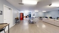 Located at 2333 Poshard Dr in Columbus, Indiana, this single-floor community game room offers a well-rounded recreational space designed for socializing and leisure. The open layout is finished with light wood-look flooring and soft gray walls, creating a clean and welcoming atmosphere throughout.

At the heart of the space sits a full-size JOOLA ping-pong table, providing a central activity for guests. Adjacent to the game area, a comfortable lounge zone features a neutral-toned sectional sofa, accent chairs, and a large wall-mounted flat-screen television — ideal for gathering and relaxing. A patterned area rug anchors the seating arrangement and adds a touch of warmth to the room.

The space also includes a dedicated dining and meeting area with a rectangular table and modern multi-chair seating, suitable for casual meals or group activities. A pair of bistro-height tables with bar stools offer additional seating options. A kitchenette with cabinetry and a refrigerator provides convenient refreshment access, complemented by a Coca-Cola vending machine positioned nearby.

Fluorescent flush lighting illuminates the space evenly, while multiple entry points — including a glass-paneled exterior door — allow natural light to filter in. The overall design balances functionality with comfort, making it a practical and enjoyable amenity for residents and visitors alike.