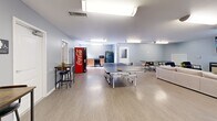 Located at 2333 Poshard Dr in Columbus, Indiana, this single-floor community game room offers a well-rounded recreational space designed for socializing and leisure. The open layout is finished with light wood-look flooring and soft gray walls, creating a clean and welcoming atmosphere throughout.

At the heart of the space sits a full-size JOOLA ping-pong table, providing a central activity for guests. Adjacent to the game area, a comfortable lounge zone features a neutral-toned sectional sofa, accent chairs, and a large wall-mounted flat-screen television — ideal for gathering and relaxing. A patterned area rug anchors the seating arrangement and adds a touch of warmth to the room.

The space also includes a dedicated dining and meeting area with a rectangular table and modern multi-chair seating, suitable for casual meals or group activities. A pair of bistro-height tables with bar stools offer additional seating options. A kitchenette with cabinetry and a refrigerator provides convenient refreshment access, complemented by a Coca-Cola vending machine positioned nearby.

Fluorescent flush lighting illuminates the space evenly, while multiple entry points — including a glass-paneled exterior door — allow natural light to filter in. The overall design balances functionality with comfort, making it a practical and enjoyable amenity for residents and visitors alike.
