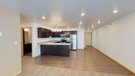 This single-floor residence offers a thoughtfully designed open-concept layout that connects the kitchen and living areas in a cohesive, functional space. The kitchen is a standout feature, showcasing rich dark cabinetry that contrasts beautifully with light granite-style countertops. A peninsula with seating overhang serves as both a prep area and a casual dining spot, while stainless steel appliances — including a refrigerator, range, and microwave — provide a modern, polished aesthetic. Recessed lighting and a stylish pendant chandelier above the peninsula ensure the kitchen is well-lit and inviting. Durable hardwood-style flooring runs throughout the kitchen and main living area, transitioning to plush carpet in the lounge space, adding warmth and comfort underfoot. Neutral white walls keep the interior feeling bright and open, allowing the dark cabinetry and wood tones to stand out as design anchors. The open flow between the kitchen and living room makes the space ideal for entertaining or everyday living. Additional doorways hint at further rooms beyond, completing this well-appointed, move-in-ready residence.