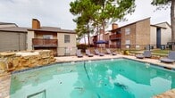 Located at Park Springs Circle in Arlington, Texas, this outdoor swimming pool area offers a well-appointed recreational retreat designed for relaxation and leisure. The centerpiece is a generously sized swimming pool with clear turquoise water, surrounded by a wide concrete pool deck that provides ample space for movement and sunbathing. A row of cushioned chaise lounges lines the poolside, offering comfortable spots to unwind, while blue market umbrellas provide shade throughout the deck.

A standout feature of this space is the natural stone water feature and adjacent spa, which add a resort-like aesthetic to the environment. The stonework is carried through in raised planters near the adjacent building, where vibrant red flowering plants add a bold pop of color against the neutral tones of the structure's lap siding and large white-framed windows.

At one end of the pool deck, a handsome wood pergola shelters an outdoor kitchen and grilling station, complete with a stone surround and dining furniture — ideal for gatherings and outdoor entertaining. The entire pool area is enclosed by a black wrought iron fence, ensuring both security and visibility. Tall pine trees frame the perimeter, providing natural shade and a lush, green backdrop that enhances the overall ambiance of this outdoor amenity space.