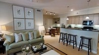 This single-floor residence features an open-concept layout that seamlessly connects the living, dining, and kitchen areas into one cohesive, well-lit space. The living area is anchored by a sage green sofa accented with decorative pillows, complemented by a glass-top coffee table adorned with tasteful décor. Botanical prints in matching frames add a refined, nature-inspired touch to the walls. Adjacent to the living area, a casual dining nook accommodates a round table with wicker-style chairs, bathed in warm light from a combination of track lighting and recessed fixtures. The kitchen features dark countertops contrasting against light wood cabinetry, with a breakfast bar equipped with sleek black bar stools providing additional seating and a natural divide between the kitchen and living space. A pendant light hangs above the bar, adding ambient warmth. Built-in shelving near the kitchen offers display space. Throughout, light-toned flooring, crown moulding, and neutral warm walls contribute to an inviting, elegant atmosphere that balances comfort with functionality.
