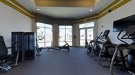 This fitness center offers a functional and welcoming environment designed to support a variety of workout routines. The space is anchored by a curved wall of large floor-to-ceiling windows and a glass entry door, flooding the interior with abundant natural light and providing open views of the surrounding outdoor area, including palm trees and neighboring buildings. The spacious, open floor plan is finished with durable dark flooring suited for high-traffic athletic use, while the walls are accented with a distinctive gold and cream color scheme that adds a touch of elegance to the practical setting. Recessed lighting in a tray ceiling supplements the natural light, ensuring the space remains well-lit throughout the day. The room is equipped with a selection of cardio machines, including rowing machines, elliptical trainers, and additional exercise equipment positioned to make the most of the available space. The curved architectural design of the window wall gives the fitness center a distinctive shape that enhances its sense of openness. Overall, the space strikes a balance between functionality and design, making it an inviting destination for daily exercise.