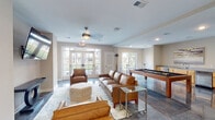 Located in Clayton, NC, this well-appointed single-floor amenity space is designed for both relaxation and entertainment. The main lounge area centers around a full-size pool table with ample surrounding space, complemented by rich leather sofas and accent chairs arranged atop a patterned area rug. A ceiling fan overhead and large French doors allow natural light to flood the room while offering views of the outdoor greenery beyond.

A built-in wet bar lines one wall, outfitted with light wood cabinetry, granite countertops, a sink, and under-counter appliances — ideal for casual gatherings. Nearby, a high-top table with bar stools provides additional seating adjacent to a window overlooking the exterior courtyard.

The TV lounge area features a wall-mounted flat-screen television, a sleek console table, and a ceiling fan, creating a cozy media space that flows naturally from the billiards zone. Polished marble-look flooring runs throughout, lending an upscale feel to the entire space.

Tucked away, a quiet dining nook offers two round metallic tables with velvet accent chairs beneath warm recessed lighting and contemporary wall art — a refined retreat within the broader communal space. Together, these zones create a versatile, modern environment well-suited for both lively recreation and relaxed socializing.