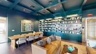 This stylish library lounge is defined by its bold, cohesive design — deep teal walls, built-in floor-to-ceiling bookshelves, and a striking coffered ceiling of the same rich hue create an immersive, curated atmosphere. The shelves are stocked with an extensive collection of books, illuminated by dedicated shelf lighting that draws the eye across every wall. The seating area centers around a pair of tufted caramel leather sofas arranged around a dark stone coffee table, accented with a decorative globe and books. A large abstract artwork adds a vibrant focal point near the dining zone. The communal dining and activity table, crafted from warm wood and surrounded by upholstered chairs, serves as a functional hub — currently home to a jigsaw puzzle in progress. Wide-plank hardwood flooring runs throughout, grounding the space with warmth. Along one wall, a full bank of floor-to-ceiling glass doors and windows floods the interior with natural light and provides direct access to an outdoor terrace with landscaped views. Recessed lighting embedded within the coffered ceiling supplements the natural light, ensuring the space is well-lit at any hour. The overall ambiance is equal parts intellectual retreat and social gathering space.
