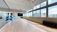 This contemporary fitness studio is designed to inspire movement and wellness in a bright, open environment. The studio's most striking feature is its expansive floor-to-ceiling windows that line an entire wall, flooding the space with natural light and offering sweeping panoramic views of a vibrant urban skyline. The light hardwood flooring throughout creates a clean, polished surface ideal for a wide variety of fitness activities and group classes. High ceilings amplify the sense of openness, and a large ceiling fan overhead ensures comfortable air circulation during workouts. A prominent large-format display screen mounted on one wall serves as a focal point for instructor-led sessions or virtual fitness programming. Adding warmth and industrial character, an exposed brick accent wall provides a striking visual contrast against the otherwise clean, modern aesthetic of white walls and recessed lighting. The studio also accommodates fitness equipment such as exercise balls, and a portable speaker stand is visible, supporting an audio-enhanced class experience. The overall ambiance is energetic yet welcoming, combining urban sophistication with a functional, well-equipped workout environment suited for group fitness, yoga, pilates, and more.