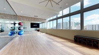 This contemporary fitness studio is designed to inspire movement and wellness in a bright, open environment. The studio's most striking feature is its expansive floor-to-ceiling windows that line an entire wall, flooding the space with natural light and offering sweeping panoramic views of a vibrant urban skyline. The light hardwood flooring throughout creates a clean, polished surface ideal for a wide variety of fitness activities and group classes. High ceilings amplify the sense of openness, and a large ceiling fan overhead ensures comfortable air circulation during workouts. A prominent large-format display screen mounted on one wall serves as a focal point for instructor-led sessions or virtual fitness programming. Adding warmth and industrial character, an exposed brick accent wall provides a striking visual contrast against the otherwise clean, modern aesthetic of white walls and recessed lighting. The studio also accommodates fitness equipment such as exercise balls, and a portable speaker stand is visible, supporting an audio-enhanced class experience. The overall ambiance is energetic yet welcoming, combining urban sophistication with a functional, well-equipped workout environment suited for group fitness, yoga, pilates, and more.