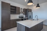 This contemporary single-floor residence showcases a clean, modern design characterized by light wood-tone flooring, crisp white walls, and minimalist finishes throughout. The open-concept kitchen serves as a focal point, featuring flat-front cabinetry in a warm taupe wood grain, a generous center island with a white quartz countertop and undermount sink, stainless steel appliances including a refrigerator and range, a wall-mounted range hood, and a subway tile backsplash. Stylish matte black pendant lights hang above the island, adding a contemporary accent. A stacked washer and dryer unit is conveniently integrated within the kitchen area. The living area is bathed in natural light from expansive floor-to-ceiling windows on multiple walls, offering sweeping panoramic views of the surrounding neighborhood and skyline. The bedrooms are well-proportioned, with large windows that continue the bright, airy feel, and each includes a sliding-door closet for ample storage. The two bathrooms are finished in a consistent modern palette — one features a walk-in glass shower and a floating vanity with a quartz countertop, while the other includes a full soaking tub with shower combo, both finished with brushed gold fixtures and light wood-tone flooring.