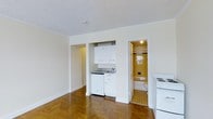 This efficiency apartment is a thoughtfully designed single-floor living space that maximizes functionality within a compact footprint. The main living area is anchored by striking herringbone-patterned hardwood floors that lend a timeless, classic character to the space. Crisp white walls and a textured ceiling keep the interior feeling bright and clean.

The built-in kitchenette is neatly integrated along one wall, featuring white upper cabinetry, a countertop with a sink, and space for essential appliances, including a stove situated conveniently nearby. The kitchen's efficient layout makes good use of every inch available. Adjacent to the kitchenette, a hallway provides additional storage or entryway space, adding to the overall functionality of the unit.

The bathroom is finished with warm yellow tile walls and a distinctive patterned tile floor, giving it a retro, vintage charm. It includes a full bathtub and toilet, providing all essential amenities. Natural light filters in through a window in the bathroom, brightening the space. Overall, this efficiency apartment offers a self-contained, practical living environment with characterful design details that make it a cozy and efficient place to call home.