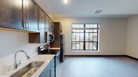 This well-designed single-floor unit offers a thoughtful layout that balances functionality and modern aesthetics. The kitchen serves as a centerpiece, showcasing rich dark-stained cabinetry that extends to the ceiling for ample storage, complemented by crisp white countertops and a full suite of stainless steel appliances, including a refrigerator, electric range, microwave, and dishwasher. Light wood-tone flooring runs throughout the main living areas, creating a warm and cohesive feel. A large picture window in the kitchen and living area invites generous natural light and frames an urban cityscape, adding an energetic backdrop to everyday living. The bathroom is clean and functional, featuring a single-sink vanity with dark wood cabinetry, a wide mirror, light countertops, and a shower with glass enclosure details. The bedroom is finished with neutral carpet flooring, soft neutral walls, and a window that allows natural light to filter in. Dark baseboards throughout the unit provide a sharp visual contrast against the light floors and walls, lending the space a polished, contemporary character. The open flow between the kitchen and living areas makes the space feel welcoming and practical for daily use.