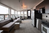 This contemporary single-floor space combines a welcoming lounge area with a fully equipped kitchenette, designed with both comfort and functionality in mind. The open layout is anchored by a generously sized sectional sofa in a soft neutral tone, accented with deep green throw pillows, and positioned to take full advantage of the space's most striking feature: a wide bank of floor-to-ceiling windows that frame sweeping views of an outdoor terrace and the open sky beyond. Natural light floods the interior throughout the day, creating an airy, inviting atmosphere. Track lighting installed on the ceiling provides additional illumination and adds a modern industrial touch. The kitchenette is sleek and well-appointed, featuring dark wood cabinetry, stainless steel appliances including a refrigerator, wall oven, microwave, and dishwasher, complemented by a stone-tiled backsplash and dark countertops. A corridor connects the main lounge to a secondary seating nook, also bathed in natural light from adjacent windows, offering a more intimate setting. Carpeted flooring in a neutral tone runs throughout, contributing to the space's warm yet polished aesthetic. Direct access to the outdoor terrace is available via a glass-paneled door, seamlessly blending indoor and outdoor living.
