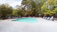 Located at 2105 Ravenglass Pl in Raleigh, NC, this outdoor pool facility offers a well-appointed recreational retreat set against a scenic backdrop of mature trees. The centerpiece is a generously sized swimming pool with clear turquoise water, surrounded by an expansive concrete sundeck that provides ample space for lounging and socializing. Multiple chaise lounge chairs and patio umbrellas are positioned throughout the deck, offering both sun and shade options for guests. Lush, tall pines and hardwoods encircle the pool area, creating a private, nature-immersed atmosphere. The facility is enclosed by a black wrought-iron safety fence, with brick accent columns adding a classic architectural touch to the entryway. A dedicated pool house structure features sliding glass doors and a separate entry door, providing access to shower and restroom facilities, in accordance with posted pool rules. Safety amenities include a 911 emergency phone station and a life ring mounted on the exterior of the pool house. The overall setting combines functional design with a serene, tree-lined environment, making it an inviting outdoor amenity for community members in Raleigh's natural surroundings.