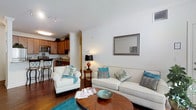 This single-floor residence offers a thoughtfully designed open-concept layout that seamlessly connects the living area and kitchen, creating a spacious and cohesive environment. The living room is a welcoming centerpiece, featuring rich hardwood floors that add warmth and character to the space. A comfortable seating arrangement, including a classic sofa and armchair accented with teal and neutral throw pillows, is anchored by a round wooden coffee table, creating an inviting atmosphere for relaxing or entertaining. A framed wall mirror and a tasteful table lamp with a decorative base add refined finishing touches. Decorative crown molding details and a painted column accent lend architectural charm to the interior. The kitchen is clearly visible from the living area, showcasing warm wood cabinetry, dark countertops, and modern appliances. A raised breakfast bar with bar stool seating provides a casual dining option while maintaining the open flow of the space. Track lighting in the kitchen and warm ambient lighting throughout the residence ensure the space is well-lit. Overall, the interior exudes a clean, polished aesthetic with a harmonious blend of classic architectural details and contemporary furnishings.