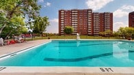 Located at 210-230 Woodridge Crescent in Ottawa's K2B neighbourhood, this outdoor amenity space serves the residents of the adjacent multi-storey brick residential buildings. The centrepiece of the grounds is a generously sized outdoor swimming pool with a surrounding concrete deck outfitted with lounge chairs, umbrellas, and colourful Adirondack-style seating, creating an inviting space for warm-weather relaxation. A lifeguard chair is positioned poolside, underscoring the facility's commitment to resident safety. The pool area is enclosed by a black iron fence and shaded by a canopy of mature trees, offering both privacy and a refreshing atmosphere. Adjacent to the pool, expansive manicured lawns provide open green space for leisure and recreation. Multiple picnic tables with umbrella shading are distributed throughout the grounds, offering comfortable spots for outdoor dining and gathering. A dedicated children's playground with colourful equipment is situated on the grounds, making the space family-friendly. A low-profile brick amenity building with informational signage anchors one section of the outdoor area, providing additional utility for residents. The entire grounds are well-maintained and secured with perimeter fencing, offering a safe and pleasant outdoor environment for all ages.