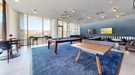 Located at 80 Bathurst Street in Toronto's vibrant King West neighbourhood, this well-appointed amenity lounge offers a dynamic and versatile social space designed for recreation and relaxation. The room is anchored by a bold, deep-blue area rug that defines the central games zone, which features both a full-size pool table and a ping-pong table — ideal for casual entertaining. Surrounding the games area, the space transitions into a comfortable lounge with tufted leather banquette seating, round tables, and upholstered armchairs arranged for intimate gatherings. The feature wall, clad in textured blue-grey brick, lends the space a sophisticated urban character, complemented by warm brass and gold accent lighting throughout. A compact kitchenette tucked along the back wall provides open shelving stocked with essentials, cabinetry, and countertop appliances. A wall-mounted flat-screen TV adds a media element to the lounge area. Floor-to-ceiling windows and sliding glass doors along the exterior wall flood the space with abundant natural light and open onto a terrace, offering sweeping views of the Toronto skyline and surrounding neighbourhood. High bar tables and stools positioned near the windows create an additional casual seating option with scenic outlooks.