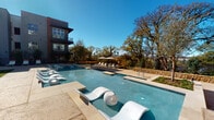 This single-floor residence offers a well-appointed interior complemented by exceptional shared outdoor amenities. The kitchen is a standout feature, showcasing dark espresso cabinetry with sleek hardware, granite countertops, subway tile backsplash, and a full suite of stainless steel appliances, including a French door refrigerator, electric range, and built-in microwave. Under-cabinet lighting adds a refined touch. The open layout connects the kitchen seamlessly to the living area, where wood-look flooring, elevated ceilings, and a teal accent wall create a warm yet contemporary atmosphere. Pendant lighting above the kitchen island adds visual interest, while natural light flows in through French doors leading to the living space. The entry foyer features panel-style doors and convenient closet access. Outside, the property boasts a resort-style pool area with a shallow sun shelf lined with sculptural white loungers, a separate spa, traditional lounge chairs with umbrellas, and a stone retaining wall. Mature trees frame the space while distant city skyline views enhance the open-air ambiance under expansive blue skies.