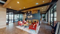 This single-floor amenity space is designed with a cohesive contemporary aesthetic that blends warm wood tones, dark slate-finished accent walls, and large-format tile flooring throughout. The main lounge area serves as the focal point, centered around a bold dark feature wall with built-in display niches finished in natural wood. A sculptural multi-arm chandelier anchors the space overhead, while a set of coral-red armchairs surrounds a reclaimed wood coffee table set with a chess board, creating an inviting social hub. Floor-to-ceiling steel-framed glass walls flood the interior with natural light and provide views of the surrounding exterior.

An adjacent secondary lounge offers a more relaxed atmosphere, furnished with a plush sectional sofa, accent chairs, and a wall-mounted television, all beneath an exposed wood beam ceiling. The space transitions seamlessly into a fully equipped kitchen and entertaining area, featuring striking dark marble countertops, a large central island with bar seating, pendant lighting, stainless steel appliances, and decorative wall art. A glass-enclosed co-working or conference room is also accessible, providing a quiet and focused environment within the broader communal space.