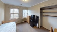 Located on South Hyde Park Boulevard in Chicago's historic Hyde Park neighborhood, this single-floor unit offers a compact and functional layout designed for efficient, comfortable living. The main living area serves as a bedroom, featuring neutral beige walls, crown molding, and wall-to-wall carpet in a neutral brown tone that runs throughout the space. The room accommodates a single bed with a wooden frame and includes a dresser, a compact refrigerator, and a microwave, providing basic in-unit convenience. A built-in closet with open shelving offers additional storage. A desk and chair are also present, making the space suitable for study or remote work. Two double-hung windows along one wall allow natural light to filter in and provide views of surrounding trees, adding a touch of greenery to the interior. A radiator beneath the windows provides heating. The private bathroom is compact and features classic penny tile flooring, a pedestal sink with a medicine cabinet mirror, a toilet, and a shower-tub combination. The unit's straightforward, no-frills design prioritizes functionality, making it a practical option in a well-established Chicago neighborhood.