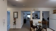 This single-floor residence at 1000 Nicholas Brooks Court in New Windsor, New York, offers a thoughtfully arranged layout that connects living, dining, and additional room spaces in a cohesive flow. The main living area features neutral white walls, carpeted flooring, and a pass-through architectural opening that provides a view into an adjacent sitting or lounge area — a distinctive design element that adds depth and openness to the space. This pass-through is framed cleanly into the wall, allowing natural sightlines and light to travel between rooms. A hallway leads to a separate room housing a television and entertainment setup, with a window that allows natural light to filter in. The dining area is positioned near the main living space and features a round glass-top table with dark wood chairs, lending a classic and functional character to the area. Decorative touches throughout — including a framed mirror, ornamental floral arrangement on a plant stand, and wall signage in the adjacent room — contribute to the warm, lived-in ambiance. The space maintains a clean, understated aesthetic throughout with consistent neutral tones and carpeted surfaces.