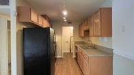 Located at 1053 Rifle Range Rd in Mt. Pleasant, South Carolina, this single-floor residence offers a well-organized and functional layout. The kitchen is designed in a classic galley style, featuring warm honey-toned wood cabinetry that extends to the upper walls, maximizing storage capacity. Upper and lower cabinets line both sides of the kitchen, providing ample space for pantry storage and cookware. The countertops offer a neutral, earthy tone that complements the cabinetry, and a stainless steel double sink is positioned conveniently along one wall. A full-size black refrigerator anchors one end of the kitchen, while a dishwasher is integrated seamlessly beneath the counter on the opposite side. Track lighting runs along the ceiling, delivering focused, adjustable illumination throughout the cooking space. The flooring throughout is a warm-toned hardwood, adding continuity and a clean aesthetic to the interior. A hallway visible from the kitchen leads to additional areas of the unit, including what appears to be a laundry connection area. The overall ambiance is practical and inviting, with a neutral color palette and quality fixtures that make this Mt. Pleasant residence a comfortable and efficient living space.