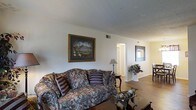 This single-story space showcases a welcoming living area characterized by a classic, traditional aesthetic. The main living space features warm hardwood flooring that extends throughout, complementing the neutral white walls and ceiling. A floral-patterned sofa with decorative accent pillows serves as the focal point of the seating area, anchoring the room with a timeless, comfortable feel.

The layout opens toward additional rooms, visible through a doorway at the rear, suggesting a practical and connected floor plan. A framed landscape painting adorns one wall, adding a touch of elegance, while a mirror on the opposite wall enhances the sense of space and reflects light throughout the room. A classic table lamp on a dark wood side table provides warm accent lighting, contributing to the inviting ambiance.

Decorative touches, including a floral arrangement on a pedestal and a small ornamental coffee table, add character to the space. Ceiling vents are visible, indicating a central HVAC system. The overall atmosphere is warm, traditional, and well-maintained, offering a comfortable and livable environment suited to classic interior sensibilities.