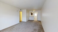 This single-floor residential space offers a clean, functional layout designed for comfortable everyday living. The main living area is finished with soft, wall-to-wall carpeting in a neutral tone, creating a warm and inviting atmosphere throughout. Crisp white walls and a textured ceiling contribute to a bright, open feel, while a ceiling-mounted light fixture provides central illumination.

A half-wall pass-through counter connects the living area to the kitchen, allowing for an open sightline between the two spaces and making the overall layout feel more expansive. A mirror is mounted above the counter, adding depth to the room. The kitchen area transitions to a tiled or hard-surface floor, providing a practical distinction between the two zones.

Natural light filters in through a windowed front door, lending the entryway a welcoming character. A second doorway leads to an additional room with warm-toned lighting, suggesting a private bedroom or utility space.

The overall ambiance is understated and move-in ready, with a neutral color palette that offers a versatile backdrop for a variety of interior styles. The straightforward single-floor layout ensures easy accessibility and a cohesive flow from room to room.