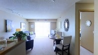This single-floor residence offers a welcoming and functional layout designed for everyday comfort. The open-concept living and dining area serves as the heart of the space, seamlessly connecting both zones for a cohesive and practical flow. The living area is carpeted throughout, providing a soft, comfortable underfoot feel, and is framed by neutral beige walls that create a warm, inviting ambiance. A large sliding glass door fills the space with an abundance of natural light, brightening the interior and offering a connection to the outdoors. Vertical blinds provide privacy and light control as needed. The dining area accommodates a round table with seating for multiple guests, positioned conveniently adjacent to the living space. A decorative sunburst mirror adorns the wall, adding a stylish focal point to the room. The ceiling features a classic textured finish, and recessed ventilation is integrated throughout. Wall art and greenery add personality and a lived-in warmth to the overall aesthetic. The layout is thoughtfully arranged to maximize usability while maintaining an open, airy feel suited to a variety of lifestyles.