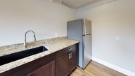 This thoughtfully designed single-floor space offers a versatile layout suited for a variety of uses. The kitchenette is outfitted with rich dark cabinetry, granite countertops, an undermount sink with a modern pull-down faucet, and a full-size stainless steel refrigerator — all set atop warm hardwood-style flooring that runs throughout the space. An open main area provides generous room to move, anchored by a striking floor-to-ceiling natural stone fireplace that serves as a dramatic focal point. Structural support columns in white add architectural character while delineating the open floor plan. Recessed lighting and a textured ceiling complete the main gathering area. A dedicated meeting or activity room features horizontal windows positioned along the upper portion of the walls, allowing natural light to filter in while maintaining privacy. The room comfortably accommodates a long table with seating, making it well-suited for group events or collaborative use. Clean white walls, consistent hardwood-style flooring, and neutral tones throughout create a cohesive and inviting atmosphere. The overall design balances practicality with character, highlighted by the standout stone fireplace and quality finishes.