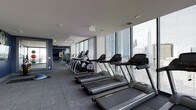This contemporary fitness center is situated on a single floor of a high-rise building, offering an impressive and well-equipped workout environment. The space is defined by expansive floor-to-ceiling windows that line the perimeter, flooding the interior with natural light and providing commanding panoramic views of the surrounding city skyline — an inspiring backdrop for any workout session.

The cardio area is prominently featured, with a row of modern treadmills positioned directly along the window line, allowing users to exercise while taking in the urban views. Alongside the treadmills, the space includes elliptical cross-trainers and additional cardio equipment, all arranged in a thoughtfully organized layout that maximizes both space and functionality.

The flooring throughout is durable rubber matting, purpose-built for a fitness environment, providing both safety and comfort underfoot. Recessed ceiling lighting complements the abundant natural light, while ceiling fans contribute to air circulation and comfort during workouts.

The overall ambiance is sleek and professional, with a contemporary design aesthetic characterized by a neutral color palette accented by deep navy tones on select walls. The combination of high-end equipment, abundant natural light, and stunning elevated city views makes this a premium fitness facility.
