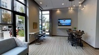 This single-floor space is designed as a contemporary lounge or common area, blending modern aesthetics with functional, flexible seating arrangements. Large floor-to-ceiling windows with dark metal framing dominate one wall, flooding the interior with natural light and offering open views of the surrounding outdoor greenery and streetscape. The expansive glazing also highlights a textured stone or wood-clad structural column, which serves as a striking architectural accent that adds warmth and visual contrast against the neutral gray walls.

The flooring consists of light-toned, wood-look plank tile laid in a linear pattern, contributing to the clean and contemporary character of the space. Seating options are varied and thoughtfully arranged: a wall-mounted counter with industrial-style bar stools sits adjacent to the windows, while a sturdy wooden communal table paired with mixed metal chairs occupies the central area, ideal for casual gatherings or collaborative work.

A large wall-mounted flat-screen television is positioned prominently on the main wall, suitable for presentations or entertainment. Pendant lighting with glass jar-style fixtures adds a stylish, curated touch overhead. Potted plants are scattered throughout, softening the modern finishes and contributing to an inviting, well-appointed atmosphere.