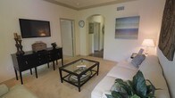 This single-story space showcases a thoughtfully designed living area that serves as the central hub of the home. The room features warm, neutral-toned walls complemented by light carpet flooring, creating a comfortable and inviting atmosphere. A distinctive arched passageway connects the living area to adjoining rooms, adding an elegant architectural detail that enhances the overall character of the space.

The living area is well-appointed, with room for a generous sofa arrangement centered around a glass-top coffee table with a dark metal frame, offering both style and functionality. A dark wood credenza along one wall provides storage and display space, while a wall-mounted television ensures modern convenience. A large abstract canvas painting adds a pop of color to the neutral palette.

Crown molding details along the ceiling add a refined finishing touch throughout. The arched hallway beyond leads to additional rooms, visible through open doorways, suggesting a practical and connected floor plan. The overall ambiance is calm and cohesive, with consistent design elements tying each area of the space together seamlessly.