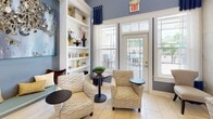 Located at 416 E North Pointe Dr in Salisbury, Maryland, this single-floor clubhouse is a thoughtfully designed community gathering space that blends comfort and sophistication. The centerpiece of the facility is a spacious, open-concept lounge area distinguished by soaring ceilings, large windows that flood the room with natural light, and a prominent white-surround fireplace with a mounted flat-screen television above it. A chandelier-style pendant light anchors the seating arrangement below, which features plush sofas, wicker accent chairs, and coordinated soft furnishings in a cool blue and neutral palette. Tile flooring throughout contributes to the bright, airy atmosphere. Adjacent to the main lounge is a well-equipped kitchen featuring dark espresso cabinetry, granite countertops, stainless steel appliances, and a tiled peninsula with bar-height seating illuminated by decorative pendant lights. A nearby casual dining area offers a round table and upholstered seating, ideal for informal gatherings. A quieter reading or lounge nook provides a more intimate retreat, appointed with patterned armchairs, a warm wood bookcase, and large windows overlooking lush greenery. A secondary seating alcove near the rear of the space opens directly to an outdoor pool area through French doors, offering seamless indoor-outdoor connectivity. Built-in shelving with curated décor accents the walls throughout, reinforcing the space's polished, welcoming character.