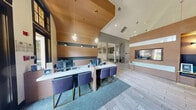 Located at 12100 Metric Blvd in Austin, Texas, this single-floor community clubhouse offers a generously sized, open-concept layout designed for social engagement and relaxation. The space is anchored by dramatic vaulted ceilings that rise prominently throughout, creating an airy, grand atmosphere.

Upon entry, a sleek marble-topped reception desk with wood-tone accents and teal carpet sets a polished, welcoming tone. Light wood-look tile flooring extends through the main corridors, complemented by warm timber slat partitions that define zones while maintaining visual openness.

The central lounge area is a bold statement in design, featuring emerald green accent walls, large arched black-framed windows that flood the space with natural light and frame views of lush greenery outside. A vibrant pink geometric-patterned carpet grounds the seating area, which is furnished with a navy tufted sofa, modern lounge chairs, and glass-topped coffee tables.

Adjacent to the lounge, a full billiards table anchors a recreation zone, while a built-in bar with blue tile backsplash, mounted flat-screen TVs, and bar stool seating provides an entertainment hub. A wood-paneled media wall with integrated lighting and a built-in bench adds further sophistication. The space also includes a fireplace and pop-art wall accents, reinforcing the eclectic, energetic character of this Austin clubhouse.