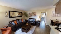 This single-floor residence offers a well-organized, open-concept layout that connects the living, dining, and kitchen areas into one cohesive space. The living room is a standout feature, anchored by rich, dark hardwood-style flooring that runs throughout the main area, providing warmth and a sense of continuity. Two dark leather sofas with coordinating accent pillows face a sturdy wooden coffee table, creating a comfortable and inviting seating arrangement. A large framed mirror and decorative wall art add character to the neutral-toned walls, while a tall indoor plant contributes a natural, refreshing element to the room. The textured popcorn ceiling adds a classic residential touch overhead. Adjacent to the living room, a dedicated dining area accommodates a dark wood table and chairs, ideal for everyday meals or entertaining. The kitchen is partially visible and features upper cabinetry in a warm wood tone, with a countertop that opens toward the dining space, enhancing the open feel. A hallway connects the main living areas to what appears to be a bedroom beyond, maintaining a practical and efficient floor plan throughout. The overall ambiance is warm, cozy, and move-in ready.