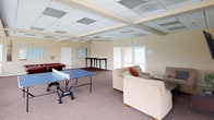This single-floor community space is thoughtfully designed to accommodate both relaxation and recreation. The open layout is unified by drop-panel ceilings with structured beam detailing, warm neutral walls, and plush carpeted flooring throughout.

The lounge area serves as a welcoming gathering spot, furnished with a sofa and armchairs arranged around a wall-mounted flat-screen television and a media console. Natural light flows generously through a wide bank of floor-to-ceiling windows fitted with semi-opaque roller shades, offering views of the outdoors including what appear to be palm trees and a sports court beyond.

Adjacent to the lounge, a dedicated game room provides ample space for active entertainment, housing both a ping-pong table and a classic billiards table. A bar-height counter area anchors one side of the room, while a comfortable sofa invites spectators to relax between games. French doors in this area offer additional access to the exterior, enhancing the sense of openness.

A separate dining area, furnished with a round table and chairs, provides a functional space for meetings or casual dining. Together, these interconnected zones create a well-rounded, amenity-rich environment suited for community gatherings and leisure activities.
