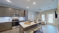 This well-appointed single-floor residence showcases a thoughtfully designed open-concept layout that seamlessly connects the kitchen, dining, and living areas. The kitchen is a standout feature, equipped with rich dark-taupe shaker-style cabinetry that extends to the ceiling for ample storage, crisp white quartz countertops, a classic subway tile backsplash, and a full suite of stainless steel appliances including a French door refrigerator, electric range with microwave, and dishwasher. A central island with bar seating serves as both a functional workspace and a casual dining hub. Light wood-tone flooring flows throughout the open living spaces, complemented by recessed lighting and a ceiling fan that enhance comfort and ambiance. The living area offers a bright, airy feel with natural light streaming through a window dressed in neutral curtains and blinds, alongside a wall-mounted flat-screen TV and access to an outdoor area. The bedroom is a tranquil retreat featuring a large mirrored closet, a bright window, and warm, neutral tones. The full bathroom is finished with large-format tile flooring, an extended dual-sink vanity with white cabinetry, backlit mirrors, and a tiled tub-shower combination, delivering a clean and modern aesthetic throughout.