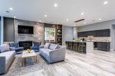 Located at 245 Atlantic Ave in Long Branch, NJ, this single-floor community space delivers a sophisticated, contemporary design throughout its open-concept layout. The interior is defined by a cohesive palette of soft steel blues, warm grays, and gold accents that carry seamlessly from one area to the next, all grounded by large-format marble-look tile flooring.

The expansive lounge area is a centerpiece of the space, featuring soaring vaulted ceilings, an abundance of natural light streaming through oversized white-trimmed windows, and a sleek floor-to-ceiling stone-clad fireplace with a wall-mounted television above. Plush gray sectional sofas and accent chairs are thoughtfully arranged around a gold-framed coffee table, creating an inviting gathering environment. Built-in shelving adds both functionality and visual interest to the room.

The kitchen and bar area is equally impressive, showcasing glossy gray cabinetry, a textured stone mosaic backsplash, gold fixtures, a beverage refrigerator, and a generous waterfall-edge island with seating for multiple guests, illuminated by a striking gold multi-pendant chandelier. A cozy bistro nook near the glass-paned entry doors offers an intimate space for smaller gatherings, completing this thoughtfully designed community amenity.