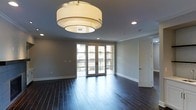 This single-floor space makes a strong first impression with its thoughtfully designed interior and quality finishes throughout. The main living area showcases rich, dark hardwood flooring that extends seamlessly across the room, grounding the space with warmth and sophistication. Neutral gray walls provide a clean, modern backdrop that complements the dark flooring and white crown molding, which adds a refined architectural detail along the ceiling's perimeter. A striking tiered drum pendant light fixture serves as a focal point overhead, offering both ambient illumination and a stylish design element, while recessed lighting throughout ensures the space is well-lit from every angle. A set of French doors floods the room with natural light and leads directly to a private balcony enclosed by iron railings, offering outdoor access and views of the surrounding area. The balcony connection enhances the sense of openness and allows for a seamless indoor-outdoor flow. The layout is open and versatile, providing ample room for a variety of furniture arrangements. Clean white baseboards and interior doors complete the polished aesthetic, making this a well-appointed space that balances classic architectural details with contemporary design sensibilities.