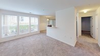 This single-floor residence offers a well-organized layout designed for comfortable everyday living. The spacious living area is finished with neutral wall-to-wall carpeting and crisp white walls, creating a clean, versatile backdrop. A large window fitted with blinds allows natural light to fill the room, while a entry door with direct access is visible from within the space.

The kitchen is designed in a galley configuration, featuring warm wood-toned cabinetry, light countertops, a stainless steel sink, and black appliances including a gas range with an overhead microwave. A peninsula with a breakfast bar separates the kitchen from the living area and is accented by a pendant light overhead. Light wood-style flooring runs throughout the kitchen, providing a practical and cohesive look.

The bathroom includes a wood-toned vanity with a single sink, a wall-mounted mirror with overhead lighting, light wood-style flooring, and a separate toilet and open shelving area for storage. The bedroom is carpeted and well-lit by windows that overlook greenery outside. A dedicated laundry area houses a full-size top-loading washer and dryer with overhead wire shelving for additional storage.