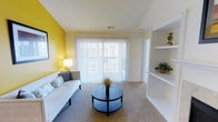 This single-floor living space is designed with a clean, functional aesthetic that balances comfort and practicality. The living area serves as the focal point of the home, distinguished by a vibrant yellow accent wall that adds a lively pop of color against the neutral white ceiling and light-toned walls. Vaulted ceilings contribute to an open, airy atmosphere, enhancing the sense of spaciousness throughout the room. Soft, wall-to-wall carpeting provides a comfortable underfoot feel and complements the room's warm, welcoming character. A set of full-height sliding glass doors, dressed with vertical blinds, allows generous natural light to fill the space while offering access to an outdoor area. The doors also provide a pleasant view outward, connecting the interior to the outside environment. Built-in white shelving units along one wall offer practical display and storage options, adding architectural interest without occupying floor space. A dedicated doorway adjacent to the sliding doors suggests access to an additional room or utility area, contributing to the overall flow of the layout. The space is well-suited for comfortable everyday living.