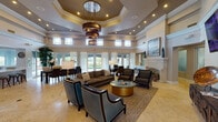 This sophisticated single-floor communal space blends upscale design with inviting functionality. The centerpiece of the main lounge is a dramatic octagonal tray ceiling adorned with a striking multi-tiered circular chandelier, lending an air of grandeur to the open-concept interior. Travertine tile flooring flows seamlessly throughout, complementing the warm neutral tones of the walls and décor.

The spacious lounge area is appointed with contemporary seating arrangements, including tufted accent chairs in rich teal and patterned upholstery, a plush sofa, and a glass-top coffee table with a gold base. A white stone fireplace serves as an elegant focal point on one wall, while a wall-mounted flat-screen television anchors the opposite side. Curated artwork and decorative accessories add refined character throughout.

A dedicated dining area features a dark wood table surrounded by upholstered chairs, bathed in natural light from floor-to-ceiling French doors draped in sheer white curtains that open to an outdoor patio. Clerestory windows positioned along the upper walls allow additional daylight to fill the interior.

The kitchen area is bright and functional, featuring white quartz countertops, grey cabinetry, a peninsula with bar seating, and a bay of windows overlooking the outdoors. Recessed lighting throughout ensures the space is well-lit at all hours, creating a polished and welcoming environment.