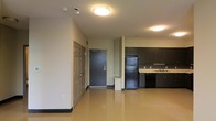 This single-floor space offers a straightforward, open-concept layout that efficiently combines living and kitchen areas. The entryway is anchored by a solid, secure front door with a keypad entry system, flanked by an interior door — offering both accessibility and privacy from the moment you arrive.

The kitchen is positioned openly along one side of the space, featuring dark espresso-toned cabinetry that extends to the upper walls for ample storage. Black appliances, including a full-size refrigerator and dishwasher, complement the dark cabinetry, while a light-colored countertop provides a clean contrast. A double sink is conveniently situated beneath upper cabinets, completing the functional workspace.

The flooring throughout is a polished, smooth surface in a warm amber tone, lending a clean and modern feel to the interior while being easy to maintain. The ceilings are fitted with flush-mount light fixtures that provide consistent, even illumination across the space.

The open plan between the kitchen and living area creates a sense of spaciousness, making the unit feel larger and highly versatile. The neutral wall tones and minimalist design create a practical, no-frills environment well-suited for a range of occupants.