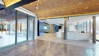 This single-floor commercial space makes an immediate architectural statement through its remarkable herringbone-patterned brick ceiling, which spans the entrance and transitions seamlessly into the open interior. The warm, textured tones of the brickwork overhead are complemented by integrated linear lighting that runs in clean, geometric lines across the ceiling, casting a refined glow throughout the space.

The flooring at the entrance mirrors the ceiling's herringbone pattern, rendered in richly toned ceramic or stone tiles that add depth and visual continuity. Large floor-to-ceiling glazed doors at the entry allow natural light to pour in, enhancing the open and welcoming atmosphere.

Moving into the interior, the space opens into a generous open-plan area defined by exposed concrete columns that lend an industrial character. Bold blue paneling and partition walls inject vibrant color, while a white reception or service counter anchors the central area. Yellow accent chairs and curved blue furnishings add playful, contemporary touches against the neutral concrete and light-toned flooring.

Glass-partitioned zones are visible in the background, suggesting dedicated meeting or collaborative areas that maintain visual connectivity with the broader space. The overall ambiance is one of modern, design-forward professionalism — balancing industrial rawness with polished, colorful contemporary interiors.
