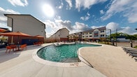 Located at 17326 N Eldridge Pkwy in Tomball, Texas, this outdoor amenity space is designed to deliver a resort-caliber experience. At the heart of the area is a generously sized freeform swimming pool with crystal-clear water, gentle water features, and a shallow wading shelf ideal for relaxing. The pool deck is finished with large-format stone pavers and lined with navy and red chaise lounges, offering ample space for sunbathing under the open Texas sky. Vibrant orange market umbrellas shade clusters of outdoor dining tables, creating a lively social atmosphere throughout the space. A covered pergola structure with warm wood beams and string lights provides an inviting shaded gathering area, complemented by a built-in brick feature for added character. The adjacent clubhouse building showcases a striking modern farmhouse aesthetic, featuring crisp white board-and-batten siding, a dark pitched roof, and expansive black-framed windows that draw in natural light. Thoughtfully landscaped grounds with ornamental trees and plantings soften the hardscape and add greenery throughout. The overall ambiance is energetic yet relaxed, blending contemporary design with comfortable outdoor living in a well-appointed community setting.