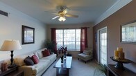 This single-floor space centers around a well-appointed living room designed for both comfort and functionality. The room features plush wall-to-wall carpeting underfoot and is finished with elegant crown molding along the ceiling perimeter, adding a refined architectural touch. A ceiling fan with an integrated light fixture is centrally mounted, providing both air circulation and ambient lighting throughout the space.

Natural light flows generously into the room through a large multi-pane window dressed with rich, deep-red curtain panels on grommet rods, which add a warm, welcoming contrast to the neutral-toned walls. A secondary door with sidelight panels and window blinds provides an additional source of natural light and outdoor access.

The living area is thoughtfully arranged with a cream-colored sofa accented by decorative red and gray pillows, complemented by a striped accent chair. A dark wood coffee table serves as the focal point of the seating arrangement. Decorative touches, including a potted plant, floral arrangement, and framed wall art, complete the inviting and cohesive aesthetic of the space.