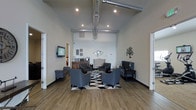 This single-floor space showcases a thoughtfully designed interior that blends industrial and contemporary aesthetics. The lounge area immediately draws attention with its impressively high ceilings, from which exposed metallic ductwork adds an industrial character against the neutral, light-toned walls. Recessed lighting is strategically placed throughout, ensuring the space is well-lit and inviting.

The seating arrangement is central to the room's design, featuring a set of slate-grey upholstered armchairs positioned around a geometric black-and-white area rug, creating a cohesive and stylish focal point. A wall-mounted flat-screen television is visible to one side, complementing the space's functionality. A beverage station or credenza is positioned along one wall, suggesting the space is designed to accommodate guests comfortably.

Decorative wall accents, including ornamental metalwork pieces, add visual interest without overwhelming the clean, minimalist aesthetic. The wood-look flooring throughout provides warmth and continuity. Access doors on either side of the main area suggest additional rooms or corridors beyond, indicating this welcoming lounge serves as a central hub within the broader space.