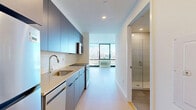 Located at 632 Newark Avenue in Jersey City, NJ, this modern studio offers a thoughtfully designed single-floor layout that maximizes every square foot. The living area is anchored by expansive floor-to-ceiling windows that frame breathtaking views of the New York City skyline, flooding the space with natural light and creating an open, airy atmosphere. Light-toned hardwood flooring runs throughout, complementing the clean, minimalist aesthetic defined by crisp white walls and recessed lighting.

The galley-style kitchen is outfitted with contemporary blue-grey cabinetry, a subway tile backsplash, stainless steel appliances including a range and microwave, and a single-basin sink — combining style with practical functionality. A hallway connects the main living areas to a dedicated laundry space and two closets, providing ample storage for a compact unit.

The bathroom is finished to a high standard, featuring large-format white tiles, a glass-enclosed walk-in shower, a wall-mounted vanity with an undermount sink, and an illuminated mirror cabinet. The overall design of the space is modern and cohesive, with neutral tones and quality finishes throughout that reflect a polished urban living experience ideally situated near the vibrant amenities of Jersey City.